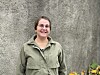 Smilende kvinne i olivengrønn jakke foran en grå vegg og blomster.