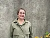 Smilende kvinne i olivengrønn jakke foran en grå vegg og blomster.