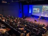 Fullsatt konferansesal med foredragsholder og presentasjonsskjerm under Blå konferanse.