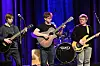 Fire musikere spiller gitar, bass og trommer på en scene med lilla og blå belysning.