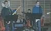 Tre musikere med gitarer og trommer spiller i et enkelt innendørs lokale.