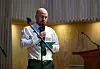 Mann i hvit skjorte taler i mikrofon foran trevegger og en talerstol.