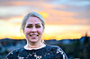 Smilende kvinne i svart blondetopp foran et uskarpt landskap ved solnedgang.