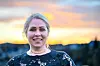 Smilende kvinne i svart blondetopp foran et uskarpt landskap ved solnedgang.