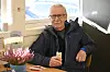 Eldre mann med briller sitter ved et bord og holder en is i et lyst lokale.