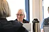 Eldre kvinne med briller ved en bærbar datamaskin, med deltakere og termos i forgrunnen.
