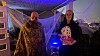 To utkledde personer står i et Halloween-dekorert inngangsparti med spindelvev, skjelett og blått lys.