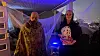 To utkledde personer står i et Halloween-dekorert inngangsparti med spindelvev, skjelett og blått lys.
